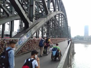 Zwei Menschen im Rollstuhl und zwei laufende Menschen ueberqueren die Bruecke ueber den Rhein bei Koeln Messe Deutz