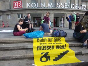 Gruppe auf dem Bahnhofsvorplatz, darunter die Bahnhofstreppe mit stufig aufgelegtem Banner 'Bahn für Alle! Die Stufen müssen weg!' Unter dem Schriftzug zeigt das Banner einen Mensch im Rollstuhl vor steiler Treppe.