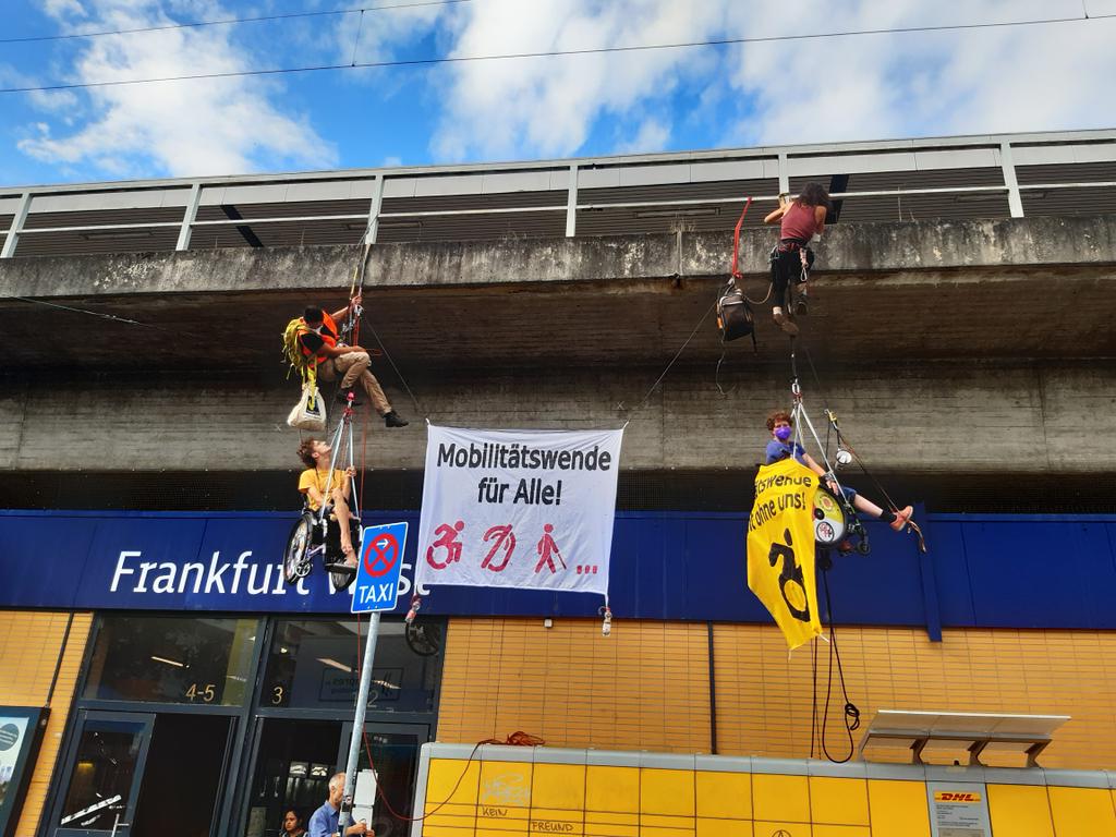 Zwei Menschen in Rollstühlen haben sich abgeseilt, dazwischen ein Transparent "Mobilitätswende für Alle" und an den Seilen auch zwei Menschen ohne Rollstühle