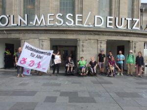 Etwa funfzehn Menschen vor dem Koeln Messe Deutz Bahnhof. Manche im Rollstuhl, manche stehen. Sie halten einen Banner wo Mobilitaetswende fuer alle draufsteht, da drunter ein symbol fuer ein Rollstuhl, ein nicht hoerendes Ohr, und ein Mensch mit Gehstock
