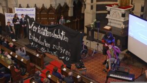 Bild mit Blick von oben (Tribune) nach unten. Bänke der Kirche mit Menschen, Leinwand mit Bühne und auf der Bühne Menschen mit einem Banner "Werkstätten abschaffen, Behinderten zuhören" und rechts davon wir einer Person im Rollstuhl von 1 Aktivisti ein Mikro gehalten.